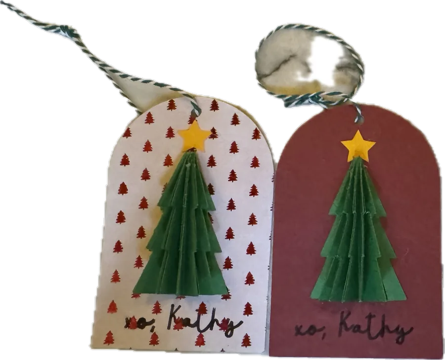 Two handmade gift tags on baker's twine, one white and one red, each with a folded paper Christmas tree, a gold star and a handwritten signature.